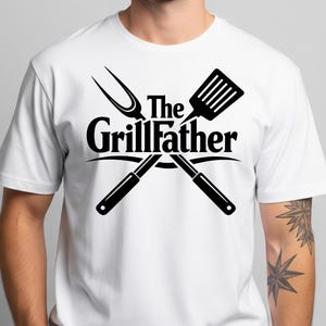 以下が含まれることがあります： 黒いグラフィックの白いTシャツ。グラフィックは、交差したグリルツールと「The Grillfather」のテキストを特徴としています。デザインにはヘラ、フォークが含まれており、テキストはツールのアーチ状になっています。