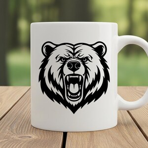 May include: White ceramic mug featuring a black and white illustration of a bear's head with an open mouth, displaying teeth. The mug has a curved handle and is set on a wooden surface with a blurred green background.