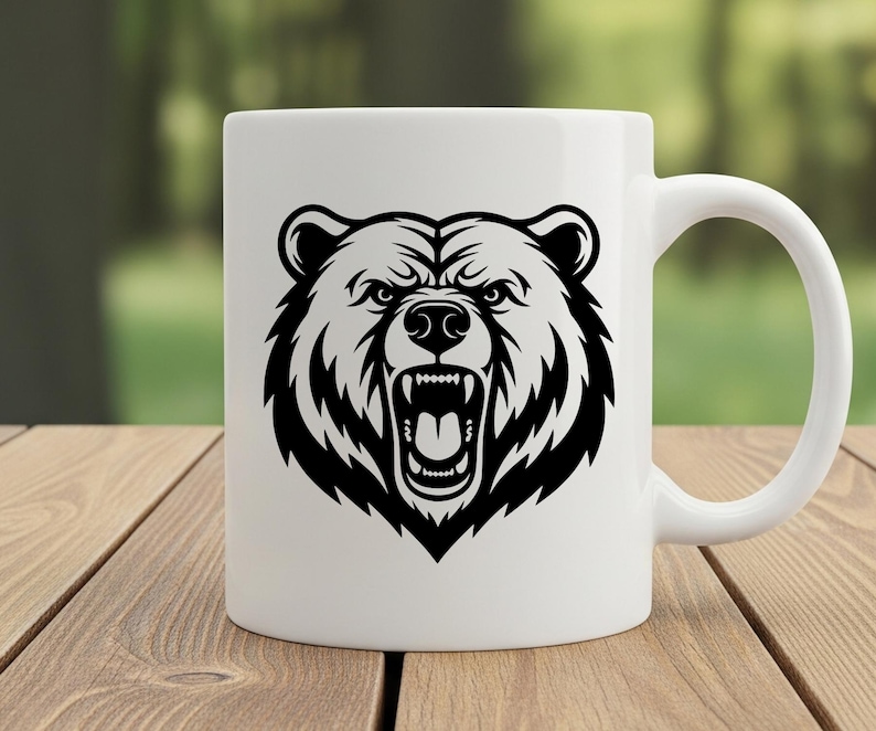 May include: White ceramic mug featuring a black and white illustration of a bear's head with an open mouth, displaying teeth. The mug has a curved handle and is set on a wooden surface with a blurred green background.