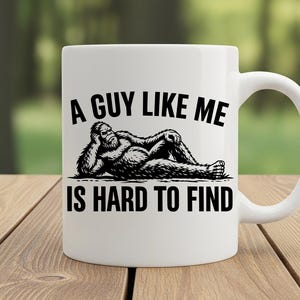 Puede incluir: Taza de cer&aacute;mica blanca con un gr&aacute;fico negro de un Bigfoot relajado y el texto "A GUY LIKE ME IS HARD TO FIND". La taza tiene un asa curva y est&aacute; sobre una superficie de madera.