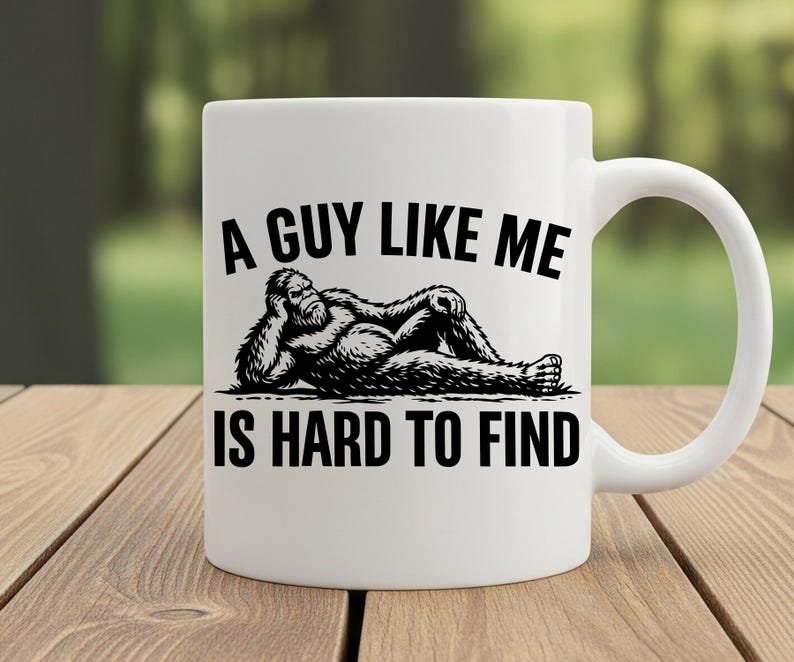 Puede incluir: Taza de cer&aacute;mica blanca con un gr&aacute;fico negro de un Bigfoot relajado y el texto "A GUY LIKE ME IS HARD TO FIND". La taza tiene un asa curva y est&aacute; sobre una superficie de madera.