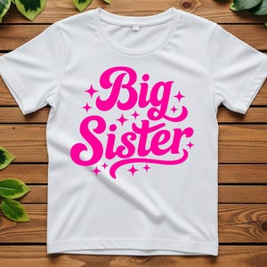 May include: White t-shirt with a crew neck and short sleeves. The shirt features the words "Big Sister" in a bright pink, stylized font, surrounded by small star shapes. The shirt is displayed on a wooden surface.
