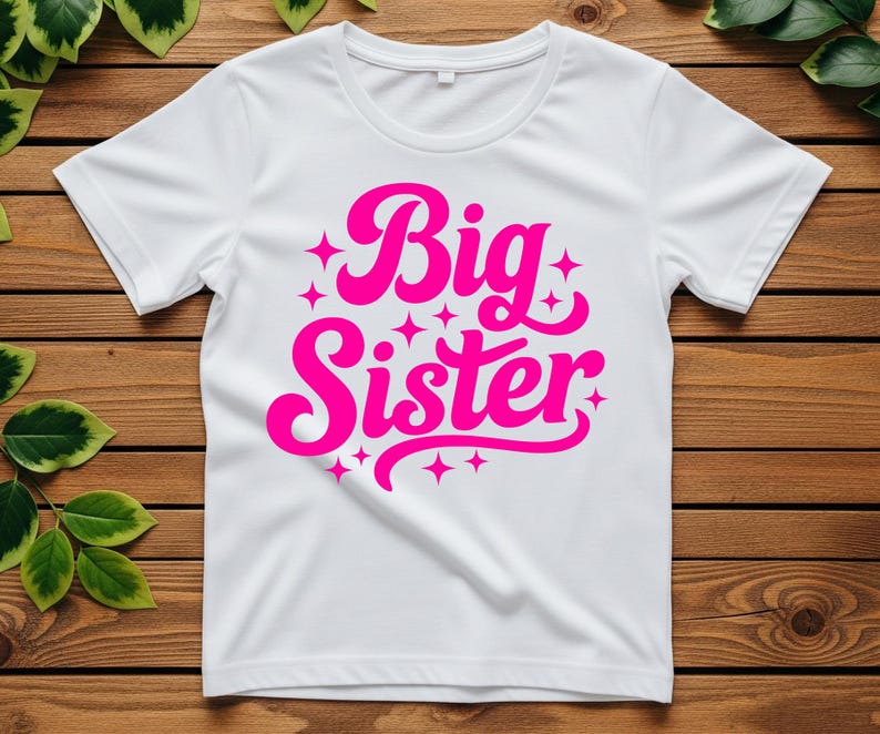 May include: White t-shirt with a crew neck and short sleeves. The shirt features the words "Big Sister" in a bright pink, stylized font, surrounded by small star shapes. The shirt is displayed on a wooden surface.