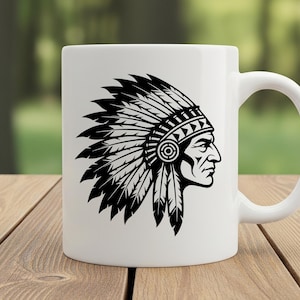 May include: White ceramic mug featuring a black and white illustration of a Native American chief in profile, wearing a feathered headdress. The mug has a curved handle and is set against a blurred green background on a wooden surface.