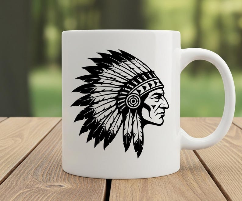 May include: White ceramic mug featuring a black and white illustration of a Native American chief in profile, wearing a feathered headdress. The mug has a curved handle and is set against a blurred green background on a wooden surface.