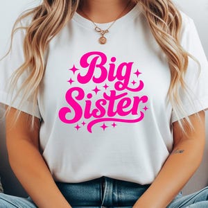 May include: White t-shirt with the words "Big Sister" in bright pink, surrounded by starbursts. The text is in a playful, cursive font. The shirt is a casual, everyday wear item.