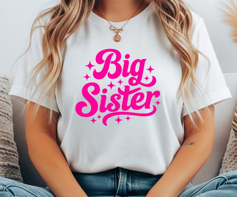 May include: White t-shirt with the words "Big Sister" in bright pink, surrounded by starbursts. The text is in a playful, cursive font. The shirt is a casual, everyday wear item.