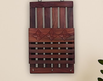 Op de afbeelding: Een donkerbruine houten wand sleutel- en posthouder. De houder heeft een lattenontwerp met gesneden bloemaccenten en kleine haken aan de onderkant. Het bovenste gedeelte heeft verticale houten latten. Het hangt aan een crèmekleurige muur.