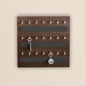 Peut inclure: Un porte-clés en bois brun foncé avec des crochets argentés et des bandes magnétiques noires. Deux jeux de clés sont suspendus aux crochets. Le porte-clés est monté sur un mur beige clair. Une tenture murale décorative est à gauche.