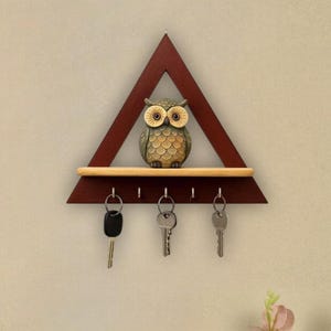Puede incluir: Un llavero triangular de madera rojo oscuro con una pequeña repisa y una figura de búho. El llavero tiene cuatro ganchos con llaves colgando. El búho es marrón y beige con ojos grandes.