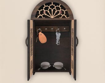 Op de afbeelding: Een donkerbruine houten sleutelhouder met een gewelfde bovenkant met een decoratief uitgesneden ontwerp. De sleutelhouder heeft een kastdeur met een sleutelrek binnenin, met twee sets sleutels en een bril.