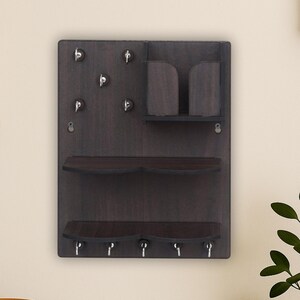 Puede incluir: Un organizador de pared de madera teñida oscura con dos estantes, ganchos para llaves y un pequeño compartimento de almacenamiento. El organizador está montado en una pared de color claro. Un cuenco decorativo con cuentas de madera y una pequeña planta en maceta están sobre una superficie de madera.