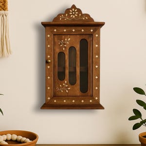 May include: A wooden wall-mounted cabinet with a dark brown finish. The cabinet features three vertical glass panels and is decorated with floral and diamond-shaped inlays. A small brass knob serves as the door handle.