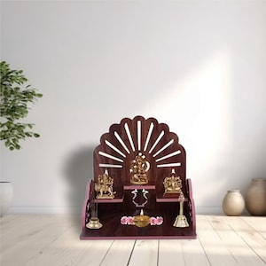 Può includere: Un altare in legno marrone scuro con una parte superiore smerlata e un design a raggi di sole ritagliati. Presenta tre statue dorate, una piccola lampada a olio e accessori decorativi in ottone. L'altare è posto su una superficie di legno chiaro.