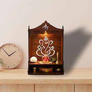 May include: A dark brown wooden temple with a cutout design of the Hindu deity Ganesha, illuminated from within. The temple features a shelf with small statues and objects. A round clock and a small plant are on the left.