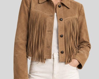 Pode incluir: Uma jaqueta de camurça castanho-claro com franjas longas no peito e nas costas, gola clássica e fecho frontal com botões circulares escuros. A jaqueta tem mangas compridas e um corte curto, usada sobre uma blusa branca e calças brancas.