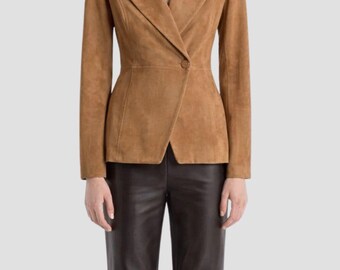 May include: A tan suede blazer with a single-button closure and a tailored fit. The blazer has a notched lapel and long sleeves. The model is wearing brown leather pants. The overall look is sophisticated and stylish.