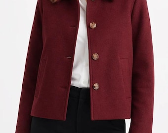 May include: A burgundy wool jacket with a collared neckline and three brown buttons. The jacket has two front pockets and long sleeves. The model is wearing a white shirt and black pants.