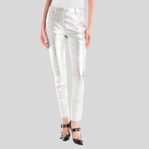May include: Silver metallic pants with a classic five-pocket design. The pants have a straight leg cut and a zipper fly. The model is wearing black high-heeled shoes with buckle details. The pants are made of a shiny, reflective material.