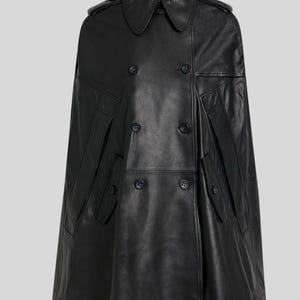 May include: A black leather cape with a double-breasted front, featuring large buttons and a classic collar. The cape has two front pockets and shoulder epaulettes. The garment is a stylish outerwear piece.