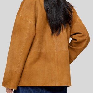 May include: A tan suede jacket with long sleeves and a boxy silhouette. The jacket has visible seams and a collar. The model is wearing a denim skirt. The jacket is a warm, neutral color.