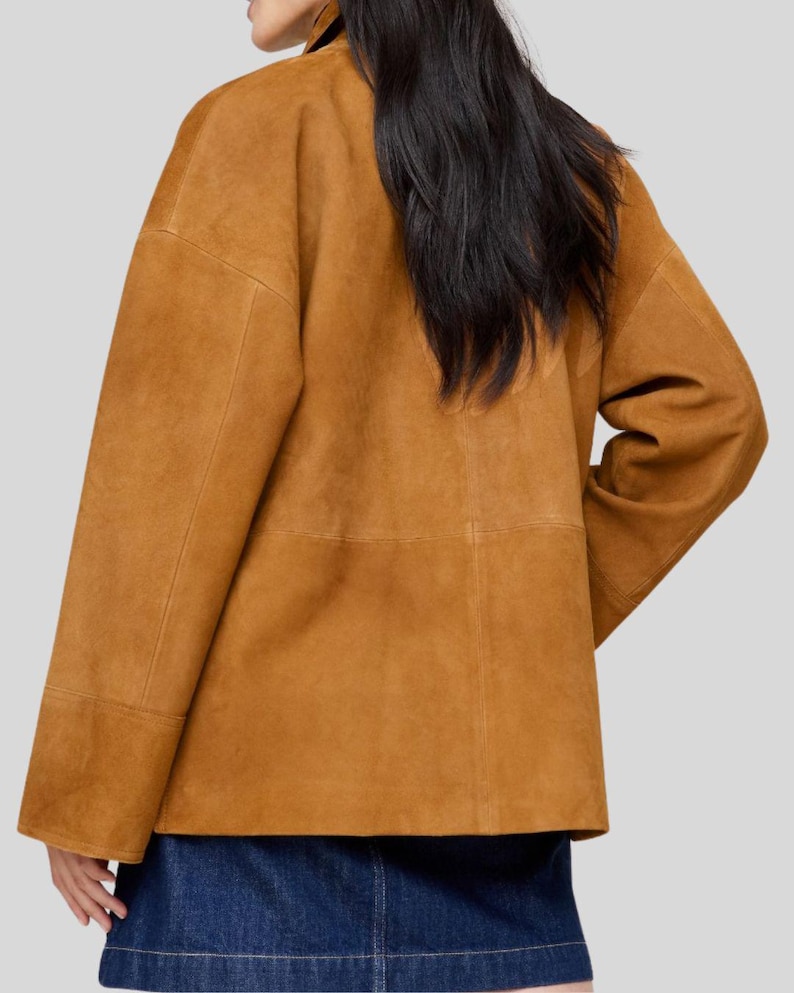 May include: A tan suede jacket with long sleeves and a boxy silhouette. The jacket has visible seams and a collar. The model is wearing a denim skirt. The jacket is a warm, neutral color.