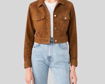May include: A brown suede jacket with two chest pockets and button closures. The jacket is worn over a white t-shirt and paired with light blue denim jeans. The jacket has a classic, cropped design.