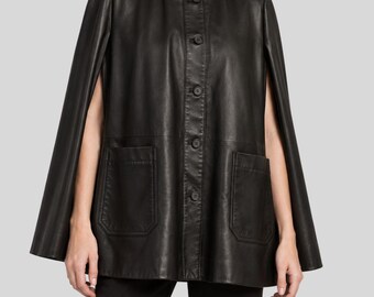 May include: A black leather cape featuring a high stand-up collar, a front button closure with five visible buttons, and two large patch pockets on the front. The garment has wide arm openings and a structured, flared silhouette that falls to the hips.