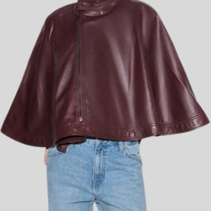 May include: A burgundy leather cape with a high collar and an asymmetrical zipper closure. The cape has a flowing, wide silhouette and is made of a smooth, shiny material. The garment is a stylish outerwear piece.