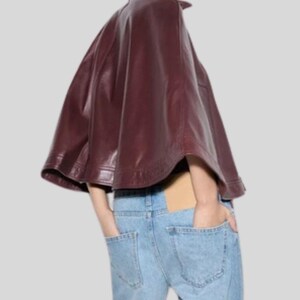 May include: A burgundy leather cape with a high collar. The cape is worn over light blue denim jeans. The model has their hands in the pockets of the jeans. The cape has a smooth, shiny texture.