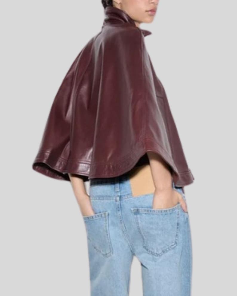 May include: A burgundy leather cape with a high collar. The cape is worn over light blue denim jeans. The model has their hands in the pockets of the jeans. The cape has a smooth, shiny texture.