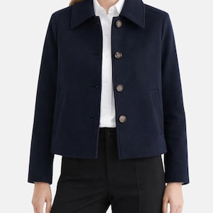 May include: A navy blue jacket with a classic collar and three brown buttons. The jacket has two front pockets and long sleeves. The model is wearing a white shirt and black pants.