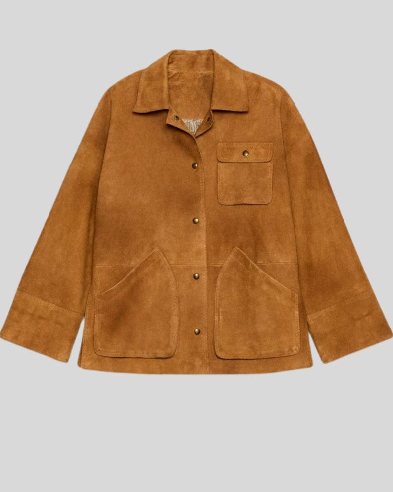 May include: A tan suede jacket with a collared neckline, button-down front, and two large front pockets. The jacket has long sleeves and a relaxed fit, perfect for layering. The jacket is a warm, earthy tone.