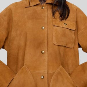 May include: A tan suede jacket with a collared neckline, button-down front, and two front pockets. The jacket has a relaxed fit and features gold-colored buttons. The overall style is casual and suitable for various occasions.
