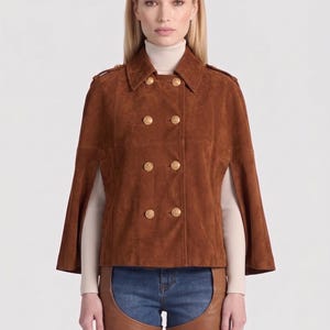 Puede incluir: Chaqueta tipo capa de ante marrón con diseño cruzado y botones dorados. La chaqueta tiene cuello y mangas cortas. La modelo lleva un cuello alto color crema y vaqueros azules.