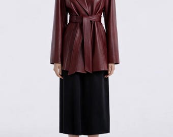 May include: A burgundy leather jacket with a belted waist and a V-neck collar. The jacket is paired with a black midi skirt and black heels. The jacket's color is a deep, rich shade of red.