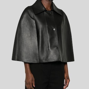 Puede incluir: Una capa de cuero negro con cuello y cierre de botones. La chaqueta tiene un dise&ntilde;o cl&aacute;sico con una textura suave. La modelo lleva pantalones negros y pendientes plateados. La capa es una opci&oacute;n de abrigo elegante.
