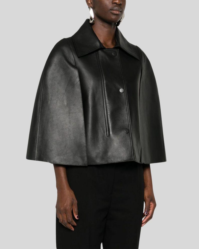 Puede incluir: Una capa de cuero negro con cuello y cierre de botones. La chaqueta tiene un dise&ntilde;o cl&aacute;sico con una textura suave. La modelo lleva pantalones negros y pendientes plateados. La capa es una opci&oacute;n de abrigo elegante.