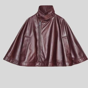 May include: A burgundy leather cape with a high collar and a front zipper. The cape features a unique design with a flared silhouette and two front pockets. The garment is made of a smooth, supple material.