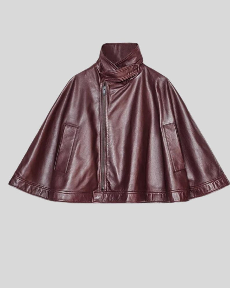 May include: A burgundy leather cape with a high collar and a front zipper. The cape features a unique design with a flared silhouette and two front pockets. The garment is made of a smooth, supple material.