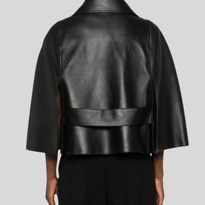 Puede incluir: Chaqueta tipo capa de cuero negro con cuello y mangas cortas. La chaqueta tiene un dise&ntilde;o estructurado con un detalle de banda horizontal en la espalda. La prenda se muestra desde atr&aacute;s.