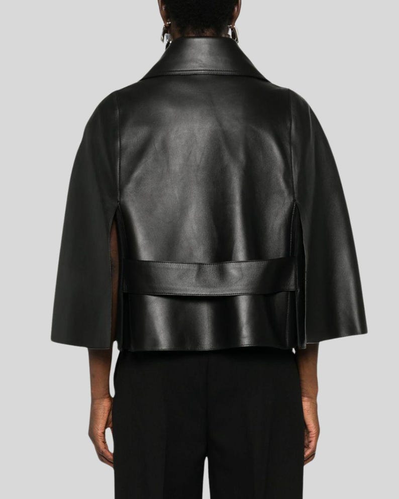 Puede incluir: Chaqueta tipo capa de cuero negro con cuello y mangas cortas. La chaqueta tiene un dise&ntilde;o estructurado con un detalle de banda horizontal en la espalda. La prenda se muestra desde atr&aacute;s.