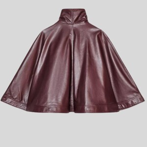 May include: A burgundy faux leather cape with a high collar. The cape has a smooth, shiny finish and a wide, flowing silhouette. The hemline is straight and the cape is designed to be worn over the shoulders.