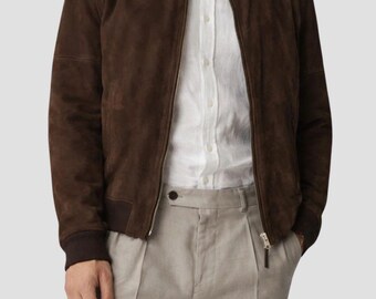 May include: A brown suede bomber jacket with a zip closure, worn over a white button-down shirt. The jacket has a ribbed collar and cuffs. The model is wearing light beige pleated trousers.