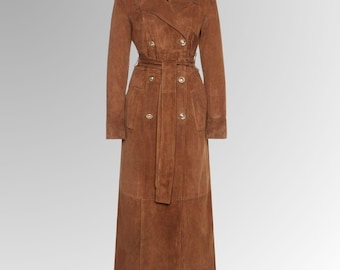May include: A long, brown suede trench coat with a double-breasted design. The coat features a belt at the waist, gold-colored buttons, and a classic collar. The coat is floor-length and has long sleeves.