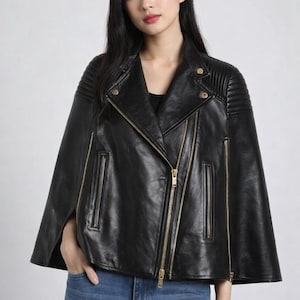 May include: Black leather cape jacket with gold zippers and accents. The jacket features a classic asymmetrical design with a notched collar and shoulder detailing. The model is wearing blue jeans.