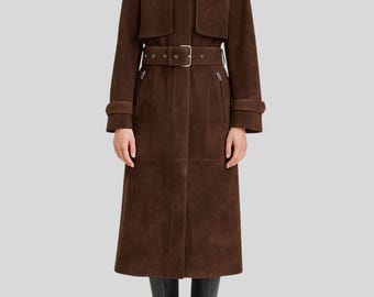 May include: A full-length brown suede trench coat with a belted waist and a high collar. The coat features a double-breasted design, a belt with a silver buckle, and long sleeves with cuff details. The model is wearing black knee-high boots.