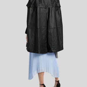 May include: A black leather cape with a structured design. The cape is paired with a light blue pleated skirt and black platform heels. The outfit is set against a light gray background.