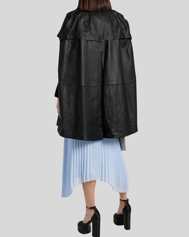 May include: A black leather cape with a structured design. The cape is paired with a light blue pleated skirt and black platform heels. The outfit is set against a light gray background.
