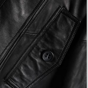 May include: Close-up of a black leather jacket featuring a buttoned pocket. The jacket has visible stitching and a textured surface. The button is dark and round, contrasting with the smooth leather.